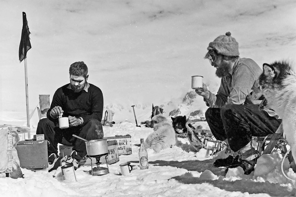 Black and white photo of two scientists making hot drinks on ice; huskies in background